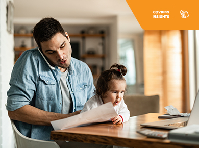 man working from home at the dining room table with toddler in his lap, phone against ear and laptop opened.