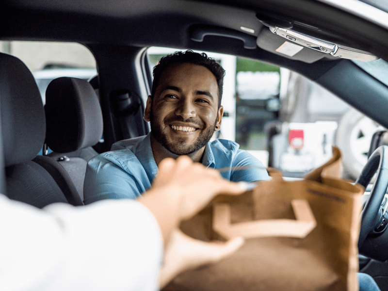 Customer in car receiving paper bagged takeout order through car window.