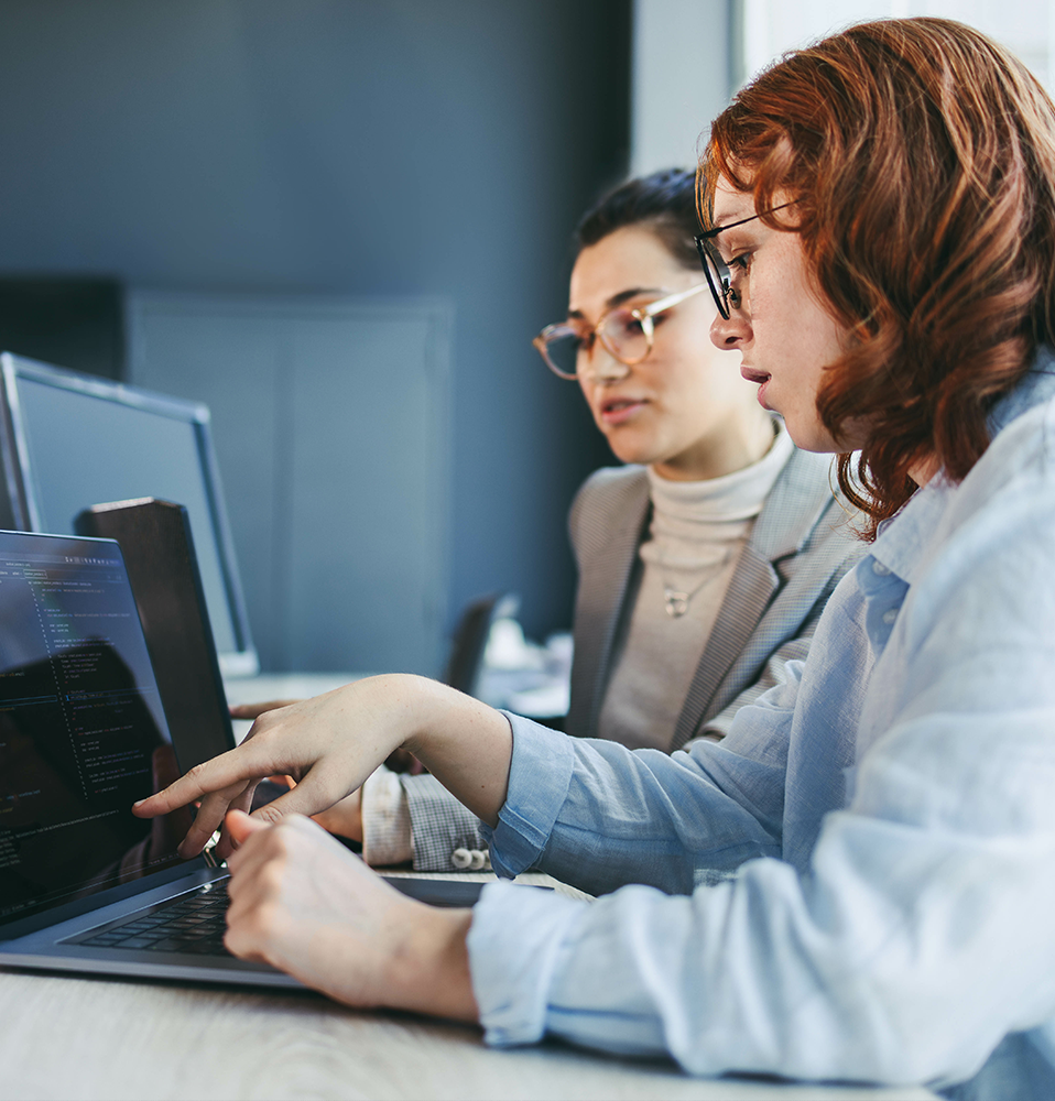 Two people collaborating on a laptop, with one person pointing at the screen displaying code.