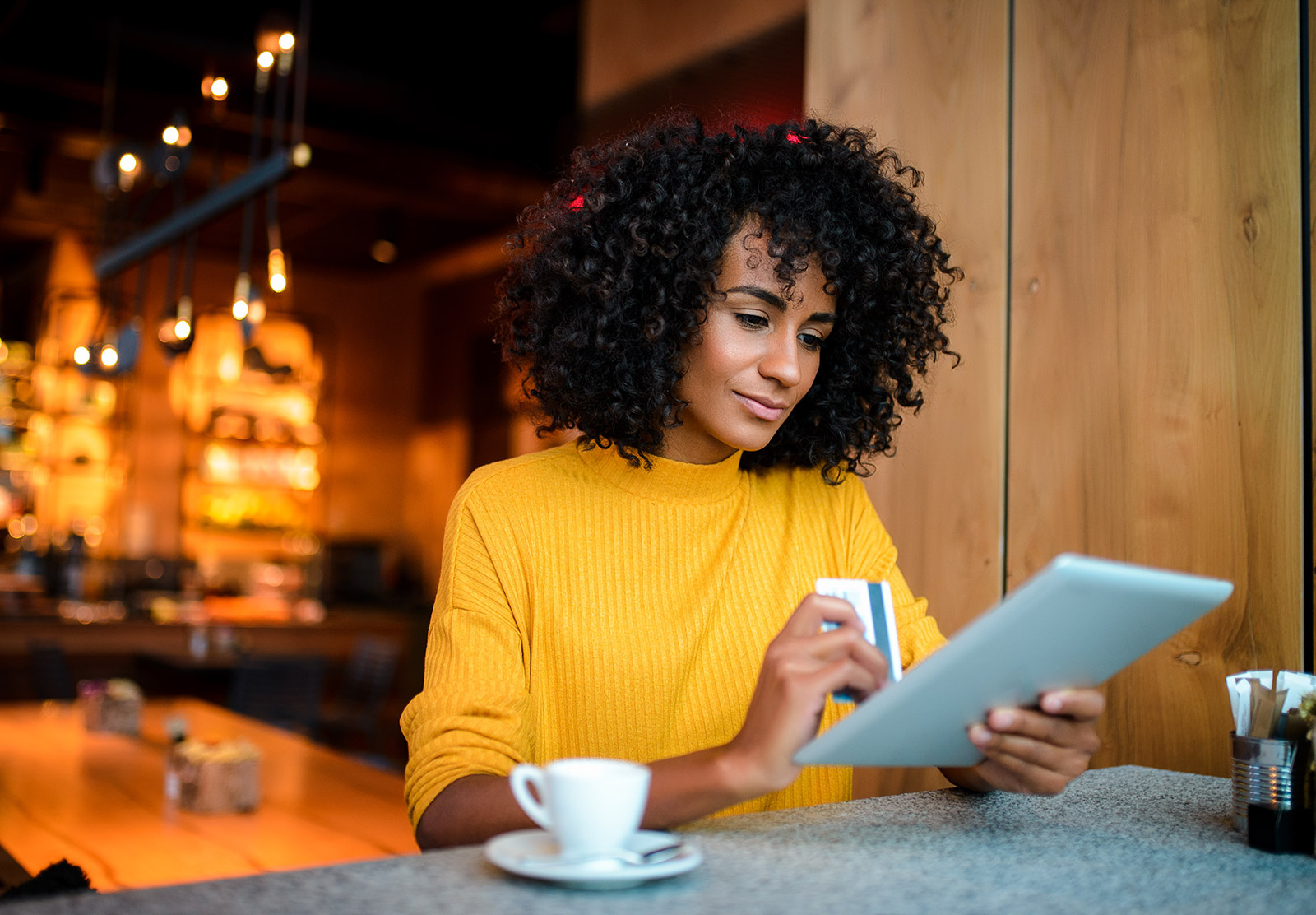 A woman holds her credit card and views personalized content on her tablet as she sits in a coffee shop