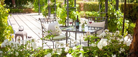  A cozy outdoor patio with a glass table set for two, surrounded by lush greenery and white flowers.