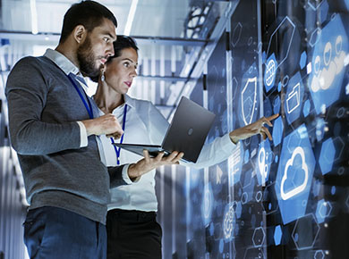 A man and woman standing with a laptop in front of a large touchscreen with icons for the hottest IT skills in 2020