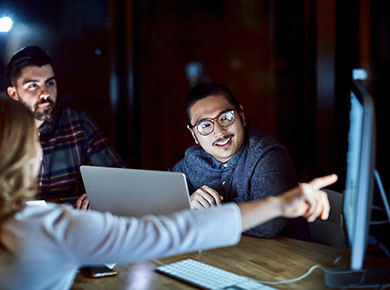 Three people sit at a meeting around a small table while one with a competitive advantage in her field points to a computer monitor