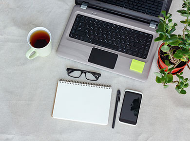 A laptop, notebook and pen, smartphone, reading glasses, cup of tea, and green plant on a white desk to help make remote working successful