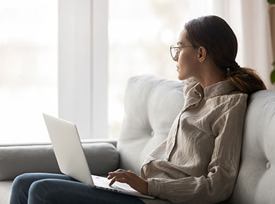 A woman sits with her laptop on a white couch by a bright window to manage wellbeing during COVID-19