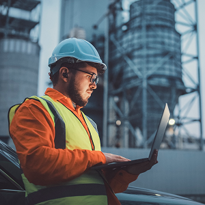 A utility worker wearing safety gear stands outdoors near industrial structures, examining a tablet device.