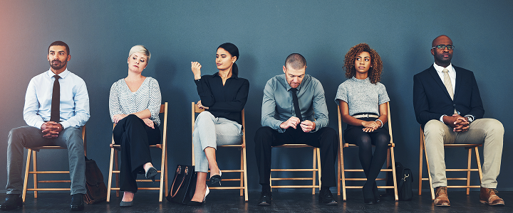 A row of people sitting on chairs in a waiting area, dressed in business and casual attire.