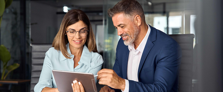 Two professionals in a modern office setting, engaged in a discussion while looking at a tablet.