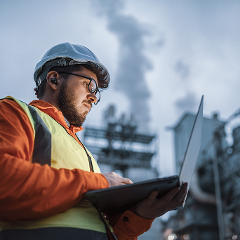 Construction worker in safety gear using a laptop at an industrial site with smoke stacks in the background.