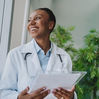 A doctor in a white coat holding a clipboard, standing near a window with green plants in the background.