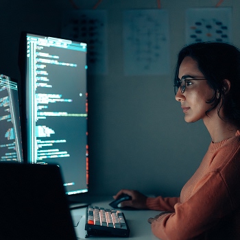 Person coding at dual monitors in dimly lit workspace with charts in background.