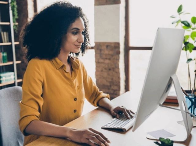 Woman in yellow button up seated in front of a computer screen as an office desk.