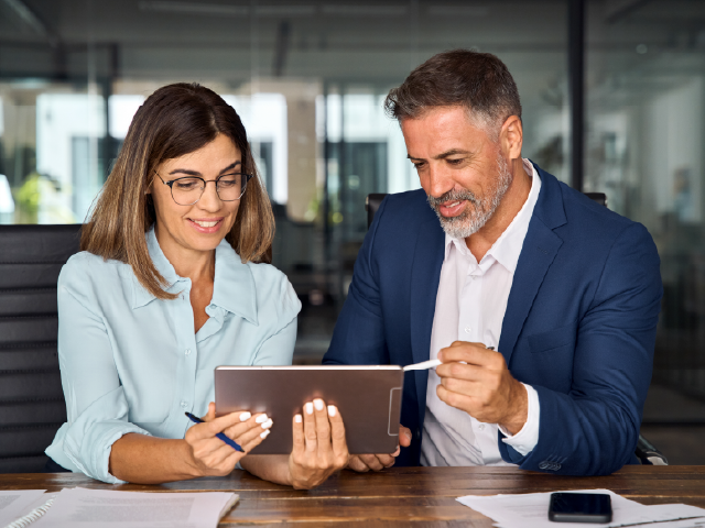 Two colleagues sitting at a desk reviewing information on a tablet together.