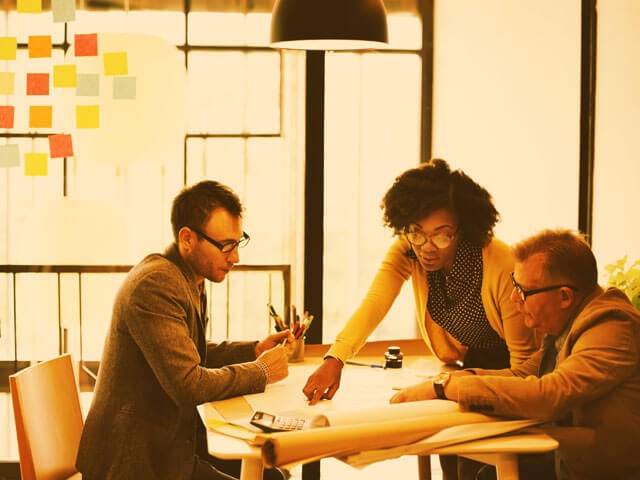three employees collaborating on a project in a conference room. Sticky notes are visible on the whiteboard behind them