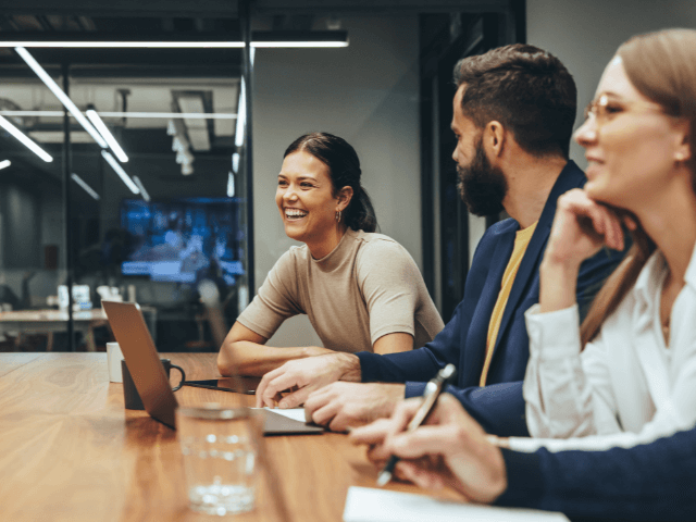a small group of young professionals smiling and talking in a conference room, seated at the table.