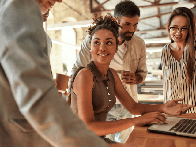 Young and cheerful afro american woman using laptop and discussing something with colleagues while sitting in the modern office.