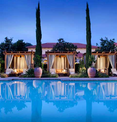 Luxurious poolside cabanas with curtains and tall potted plants, reflecting in a clear blue swimming pool at dusk.