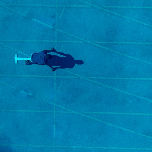 Overhead view of a lone runner moving across a running track, with lane lines and a long shadow visible.