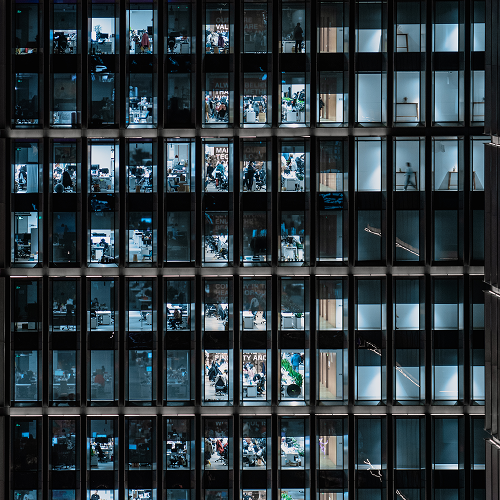 Nighttime view of a tall office building with rows of illuminated windows showing busy workspaces inside.