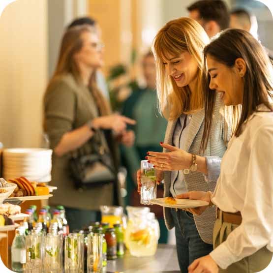 A group of people is gathered around a table with various food and drink options, engaging in conversation and serving themselves
