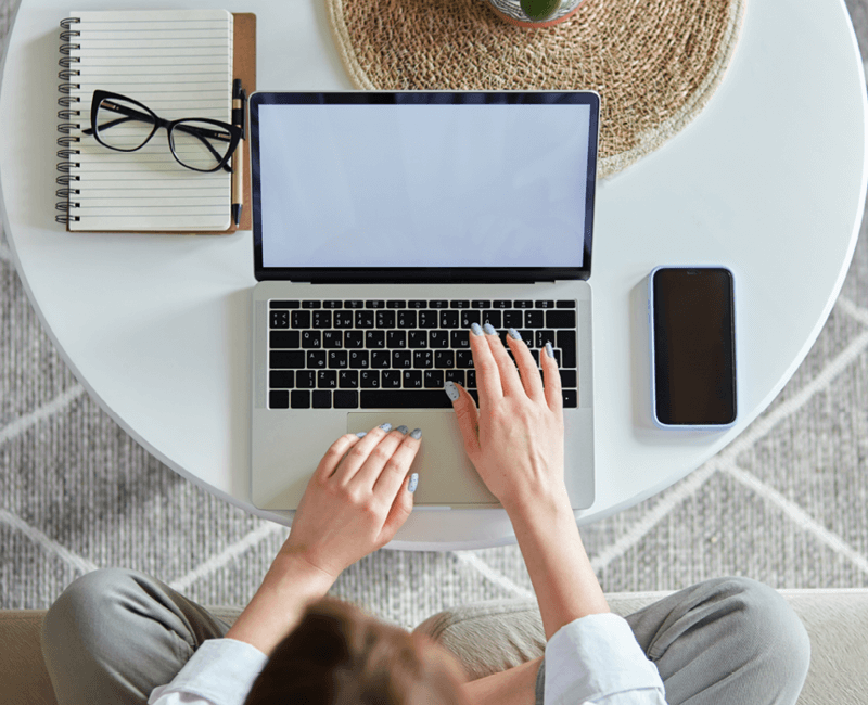 View from above of a woman seated on the floor working at a laptop on a table. Glasses and a sunhat are also visible on table.