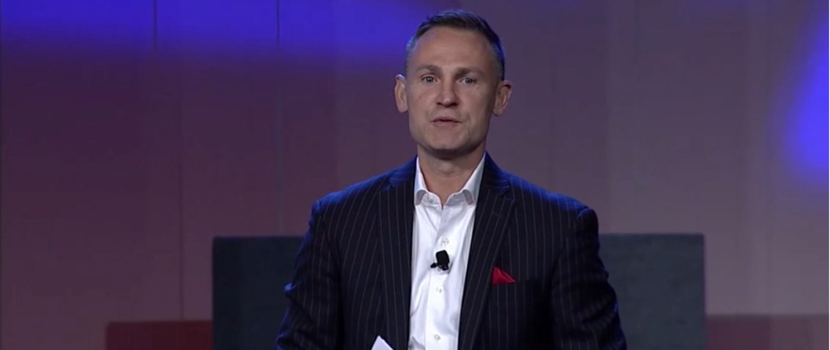 Mark Collins in a dark pinstripe suit speaking on stage with purple and blue lighting.
