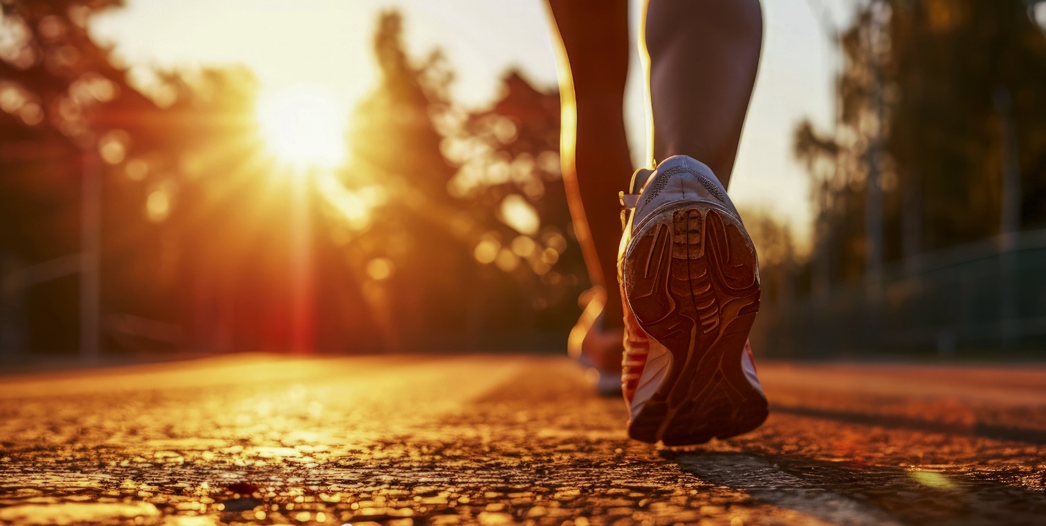 Close-up of a runner’s shoes on a paved path at sunrise, with warm sunlight illuminating the ground and trees in the background.