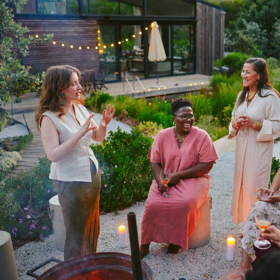 A group of four people are gathered outdoors in a garden setting, with three standing and one seated, holding drinks and surrounded by candles and string lights, near a modern house with a deck and large windows