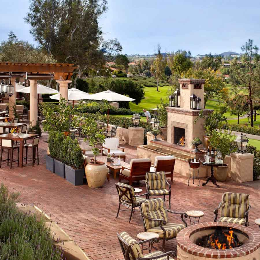 A cozy outdoor patio with cushioned seating, a fire pit, and a fireplace, surrounded by greenery and overlooking a golf course.