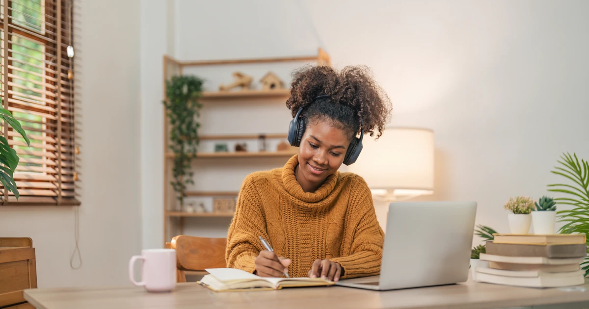 young woman in turtle neck sweater writing in a journal at a table next to a mug and open laptop