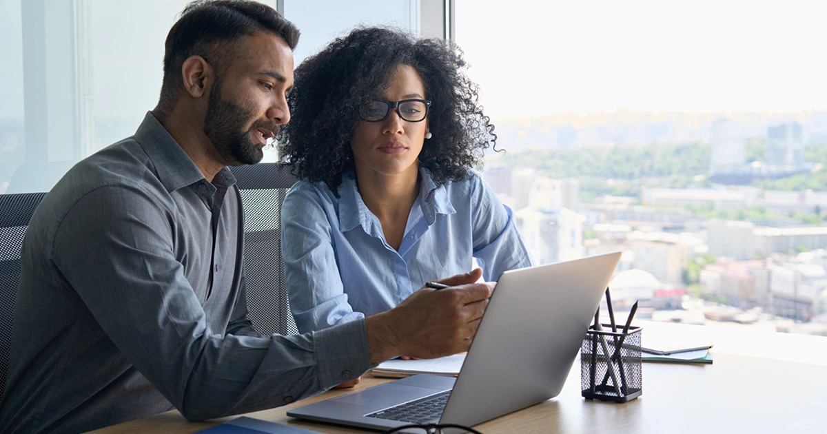 Two young professionals reviewing documents on an opened laptop seated next to a office window.