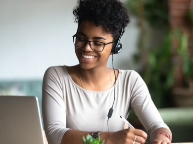 woman smiling looking at laptop screen while writing amd wearing a headset.