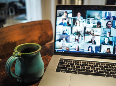 Coffee mug resting on table next to an open laptop with a video conference call displayed, multiple attendees in a grid format
