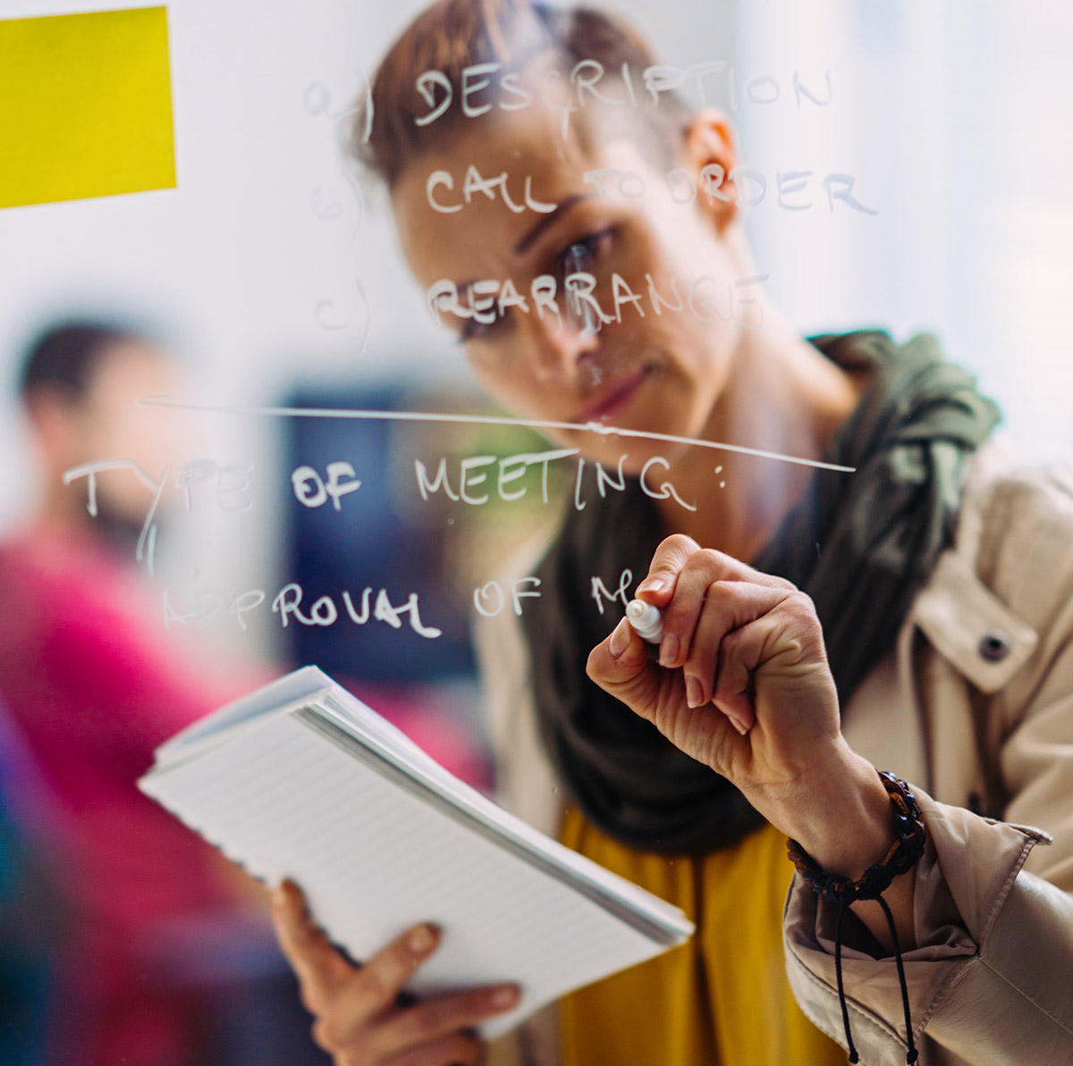 A woman in a creative studio studies a notebook as she writes on clear glass in the image foreground