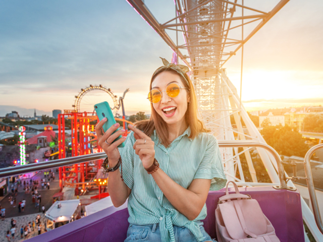A woman on a theme park ride pointing to her phone with a sunset in the background